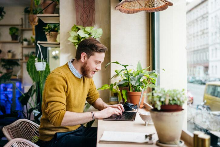 A young man sitting in a small café filled with green plants, using his laptop.