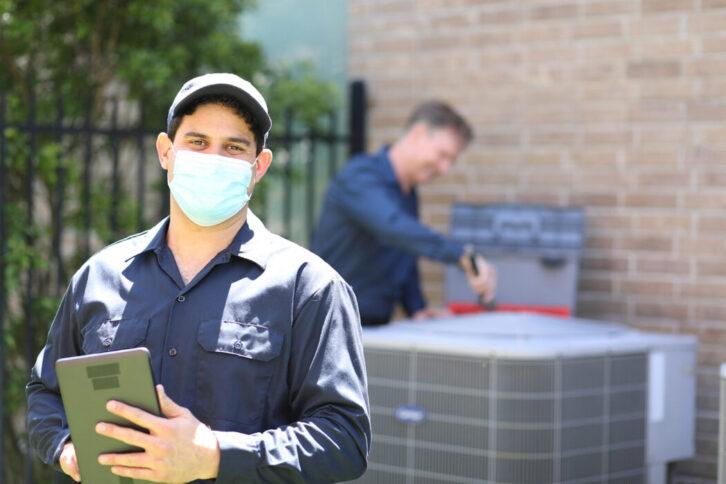 Multi-ethnic, mixed age team of blue collar air conditioner repairmen working at residential home.  They prepare to begin work by gathering appropriate tools and referring to digital tablet. Latin descent man stands out front wearing mask.