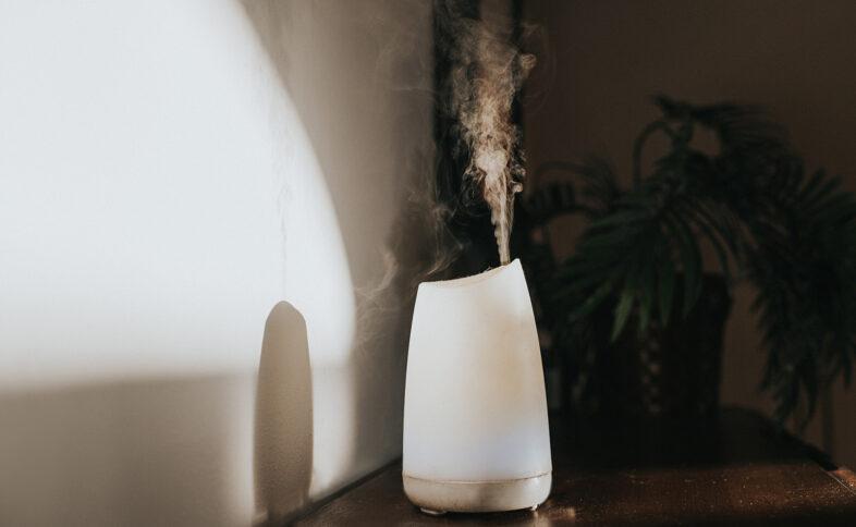 A simple white oil diffuser spritzing a light mist of water and aromatherapy blended oils into the air. It creates a shadow on the wall. Space for copy.